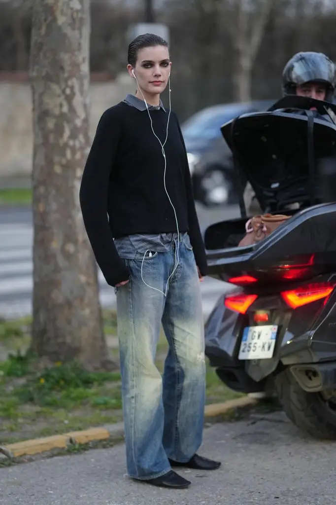Betsy Gaghan wears a gray collared shirt, black crew-neck sweater, relaxed-fit blue denim jeans, black pointed-toe leather flats, and white wired headphones. 