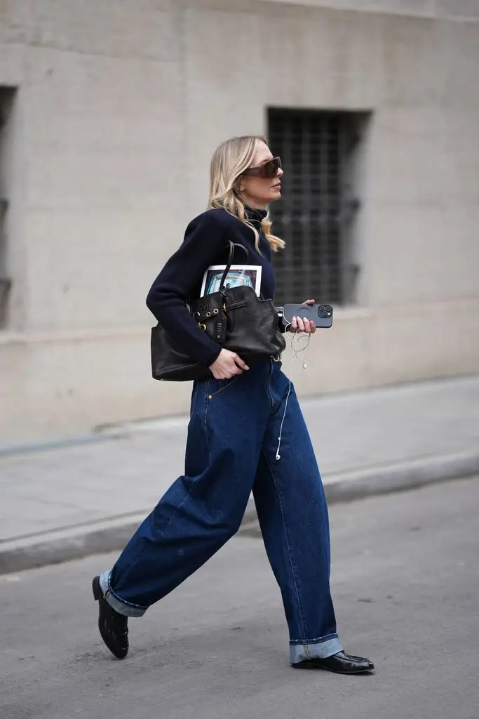 Mirja Klein wears a navy fine knit turtleneck sweater with long sleeves and a fitted silhouette from COS; dark baggy jeans with a relaxed oversized barrel fit and rolled-up legs from Dark Park; a thin black leather belt with silver buckle; black polished leather loafers with a sleek rounded toe and slim silhouette from COS, a leather bag with gold-tone details, chain link details and a belt on the front closure from Miu Miu; oversized shield sunglasses with thick clear acetate frames and brown tinted lenses from Bottega Veneta; oversized brown lens sunglasses with dark tinted lenses and thick, shiny frames from MM6 Maison Margiela; holding an iPhone smartphone and wearing white wired headphones; her blonde hair is long and wavy with a middle part 