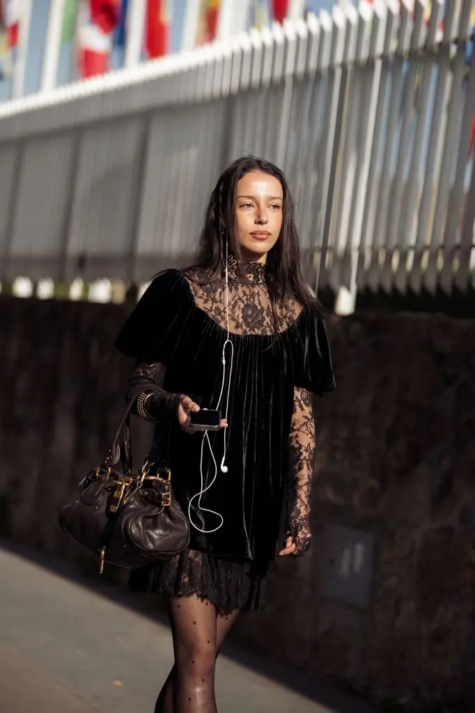 A guest wears a black velvet mini dress with puff sleeves layered over a sheer black lace long-sleeve top and lace hem, sheer black polka dot tights, a small brown leather handbag, and white embellished pointed-toe heels outside the Chloé show. 