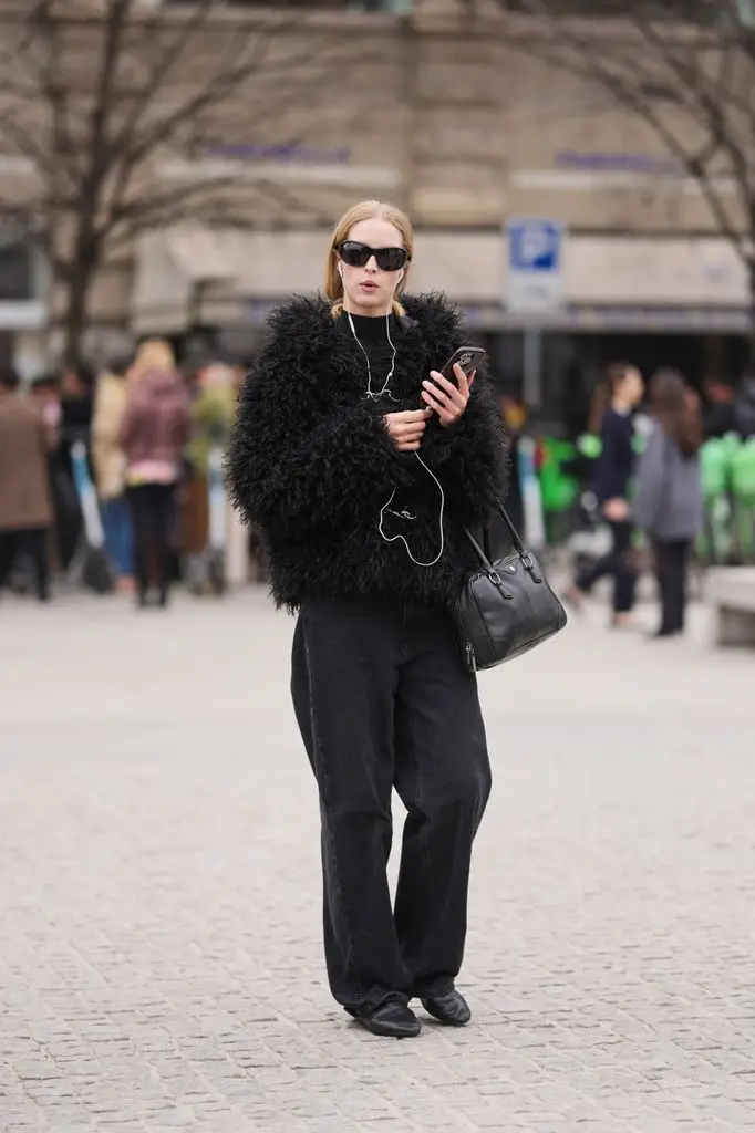 A model wears blonde hair in a ponytail, black sunglasses, wired headphones, black faux fur coat, black denim wide leg jeans, black leather bag, black leather boots.