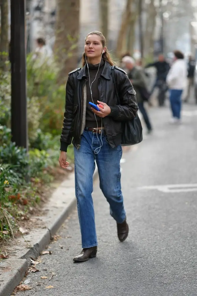 Angelina Kendall wears a dark brown leather bomber jacket, dark brown turtleneck sweater, brown leather belt, blue straight jeans and dark brown leather boots, while carrying a black leather backpack and blue phone case with white wired headphones.