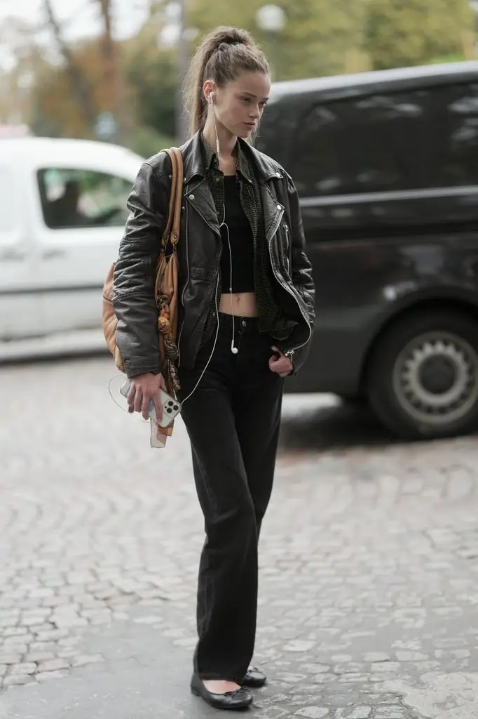 Charlotte Boggia is wearing a high ponytail hairstyle, a black leather biker jacket, a black crop top, a dark plaid button-down shirt under the jacket, black jeans, black ballet flats and carrying a beige oversized shoulder bag while holding a smartphone with white headphones. 