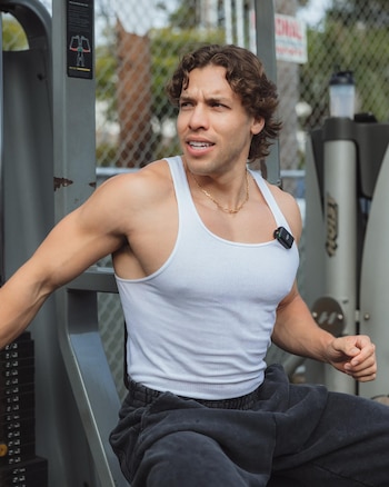 Joseph Baena, with curly hair and a white tank top, works out on an outdoor gym machine. A microphone is attached to his shoulder