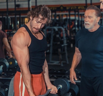 Joseph Baena, in a black T-shirt, flexes his biceps with a barbell in a gym, as Arnold Schwarzenegger looks on. dark background