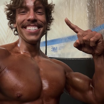 Close-up of bodybuilder Joseph Baena, with tanned and muscular skin, smiling at the camera and pointing his right index finger up