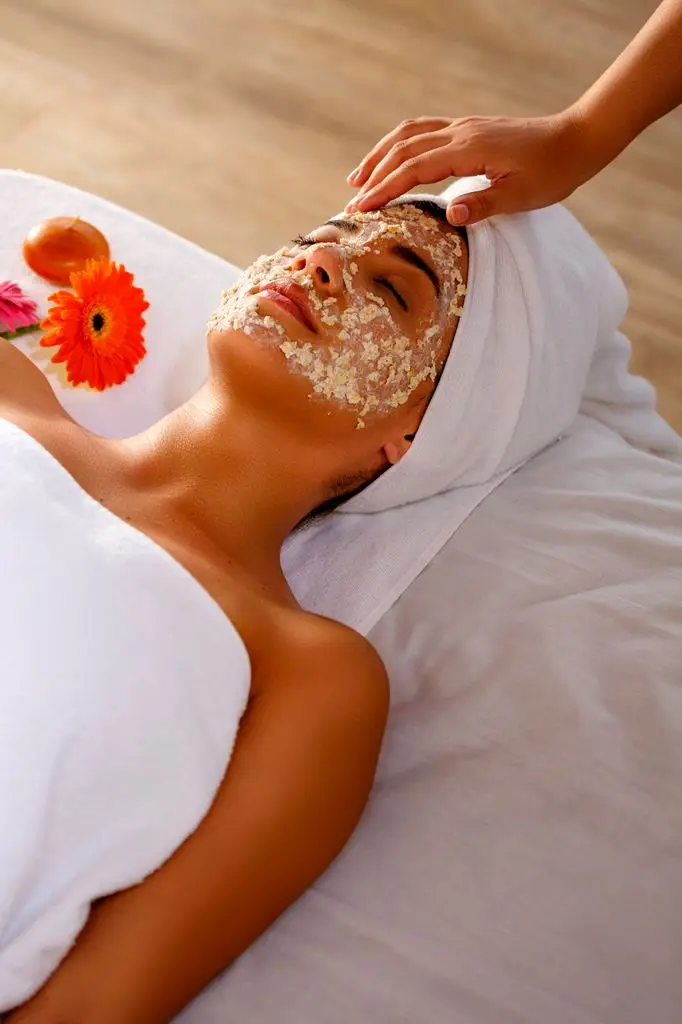 Young Hispanic woman lying on bed and scrubbing her face with oatmeal after bath