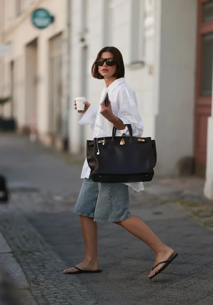 Sophia Geiss wears an oversized white structured shirt from Black Palms, loose blue-gray Bermuda shorts from Drykorn, a large Birkin, black flip-flops from Havaianas, while holding a cup of coffee and an Apple iPhone paired with wired Apple EarPods.