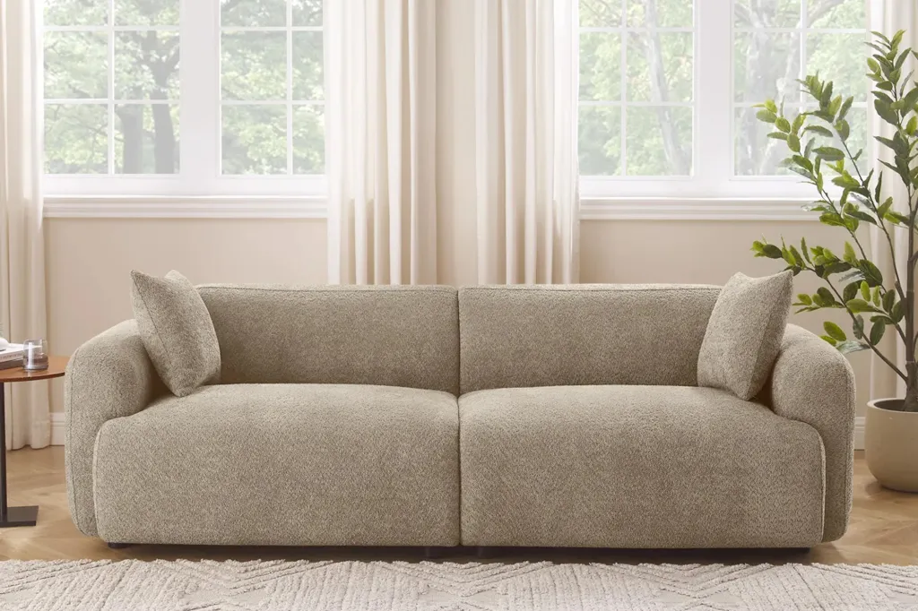 A beige textured sofa with two pillows in a living room with large windows, curtains and a plant.