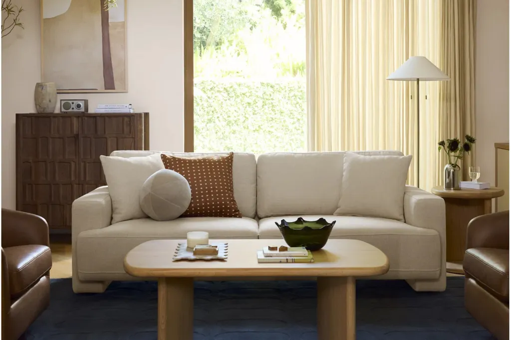 A bright living room features a cream-colored sofa with a light wood coffee table and a dark wood cabinet, with light streaming in through a large window.