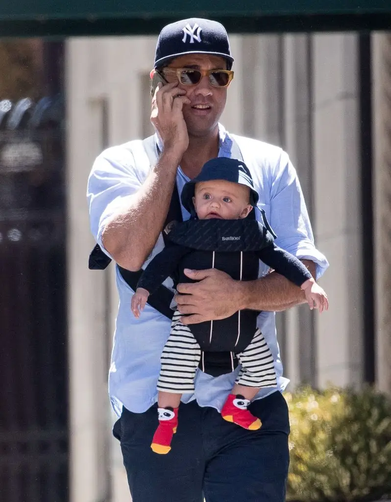 Bobby Cannavale holds his phone to his ear and holds his other arm around his son Rocco who is strapped in a baby carrier around his front.