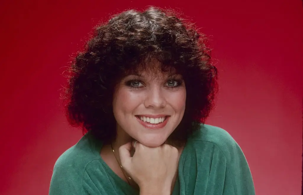 Erin Moran in a promotional photo for Happy Days wearing a green top and resting her head on her hand against a red background.