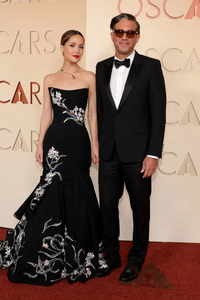 Rose Byrne and Bobby Cannavale at the Oscars in 2026. Rose wears a strapless black dress with a layered mermaid tail style and floral print. Bobby wears a tuxedo and bow tie. 