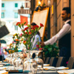 An outdoor restaurant in Italy with a floral centerpiece, empty glassware and a waiter in the background