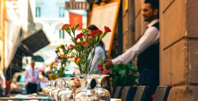 An outdoor restaurant in Italy with a floral centerpiece, empty glassware and a waiter in the background