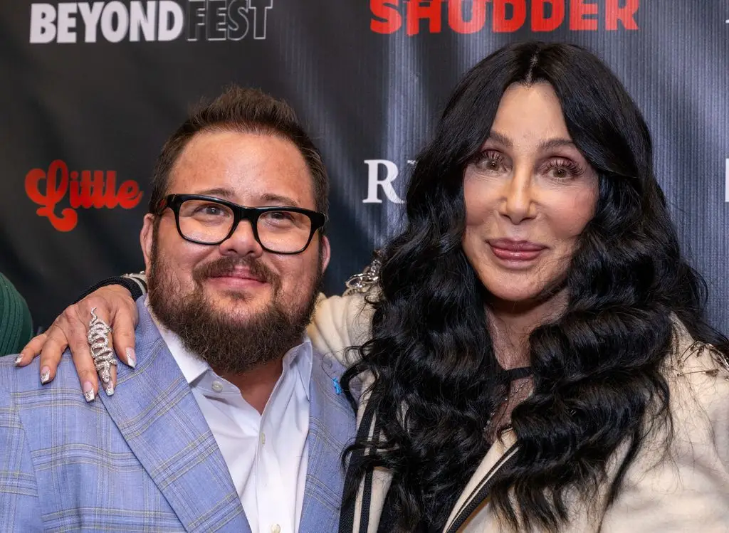 Chaz Bono in a blue suit and white shirt, standing next to his mother Cher who has an arm around her son.