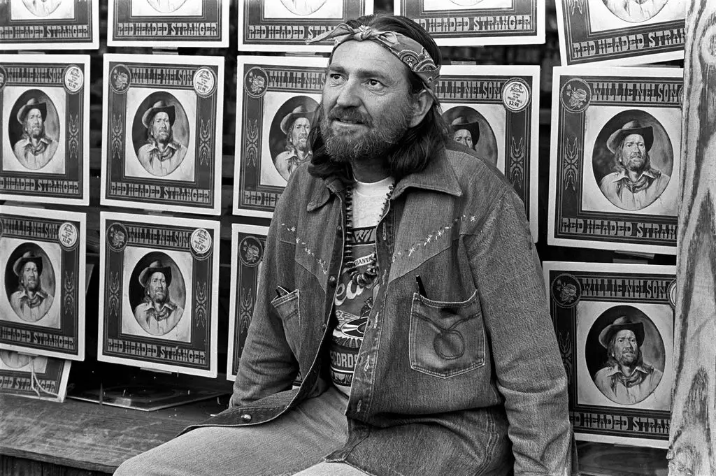Willie Nelson sits in front of several posters printed to promote his 1975 album 
