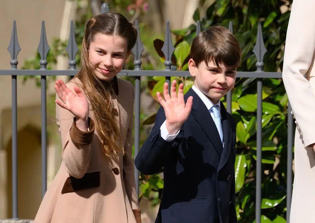 Two children, a girl in a beige coat and a boy in a black suit, greet the camera in front of a black gate.