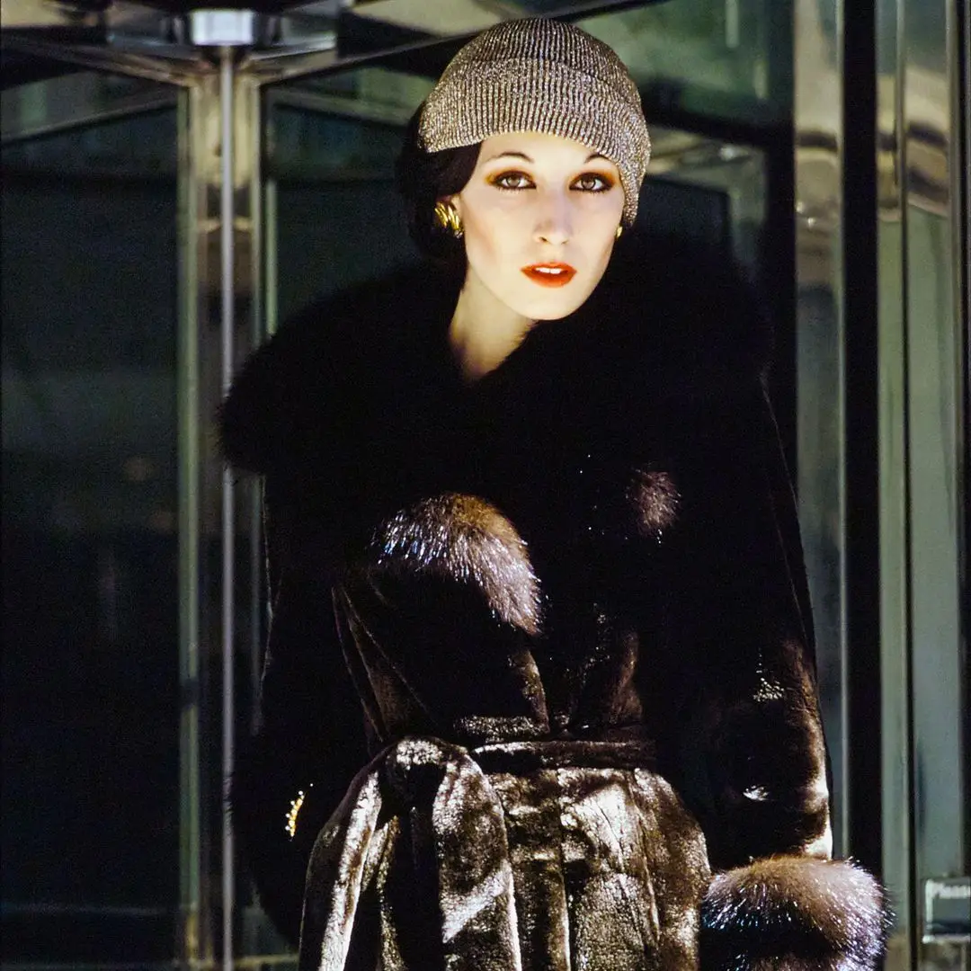 Model and actress Anjelica Huston standing in front of a revolving door wears a sealskin fur coat with sand collar and cuffs by Emeric Partos. Made from Fur Seal Safari Cape in Fouke treated silver. Lurex ribbed knit hat by Don Anderson for Scheer Brothers, Aris Gloves and Tiffany & Co. Hair jewelry by William of Kenneth. 