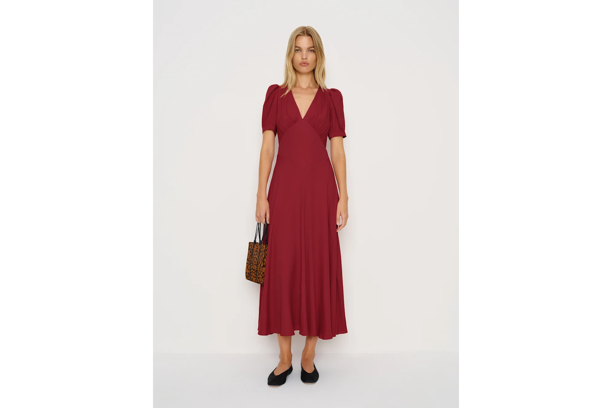 A woman wearing a long red dress with a V-neck, short puff sleeves, black flat shoes and a leopard print handbag.