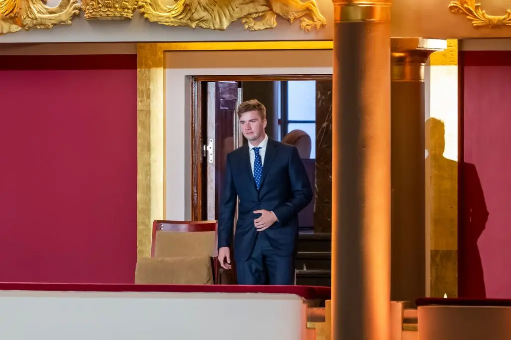 Prince Gabriel in a blue suit walks towards a chair