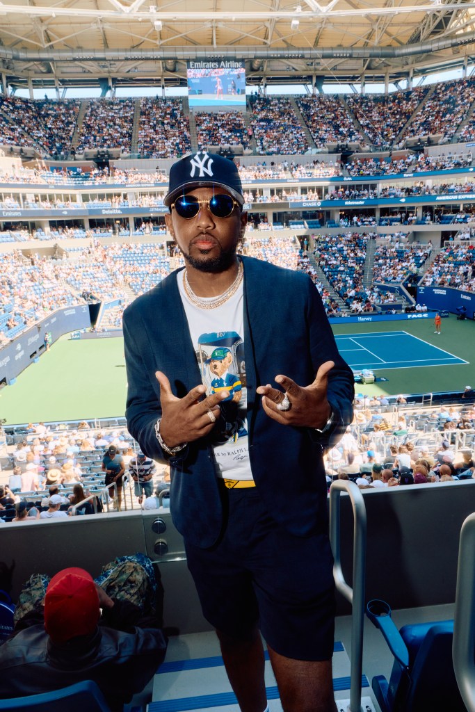 Fabolous with a Yankees cap and sunglasses poses in a stadium with a tennis court and spectators visible in the background.