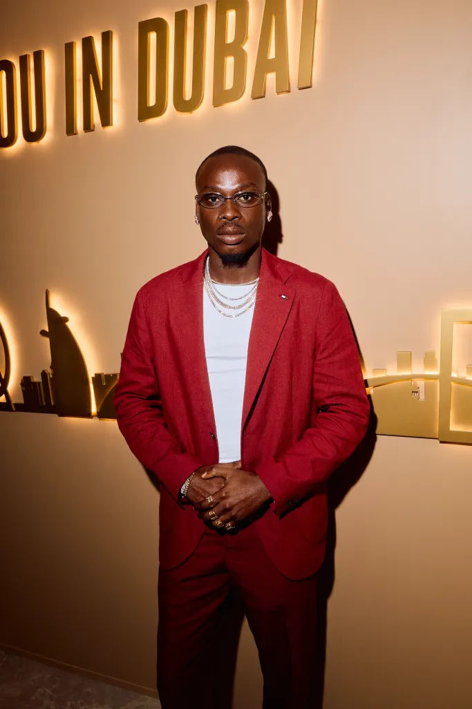 Fireboy DML in a red suit, white shirt and several chains and diamond rings posing in front of a light "DUBAI" Dubai cityscape sign and silhouette.