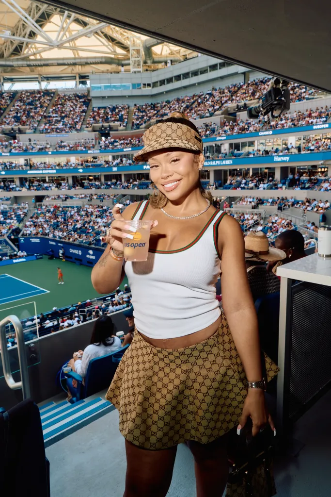 Latto wearing a white tank top, patterned Gucci visor and matching skirt poses at the US Open, holding a drink.