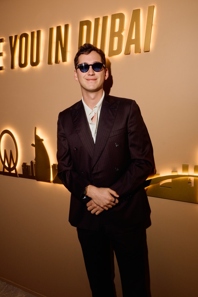 A man wearing sunglasses, a double-breasted suit and a light-colored collared shirt stands smiling in front of an illuminated sign reading "See you in Dubai."