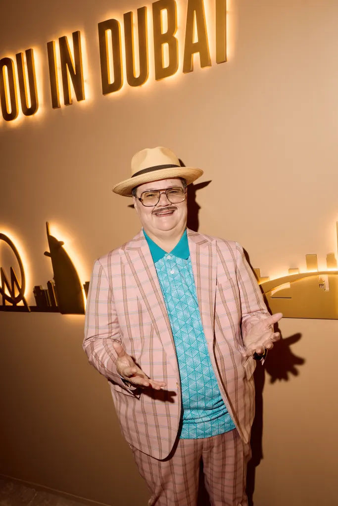 Man in plaid suit, teal shirt and fedora standing in front of lighting "DUBAI" sign.