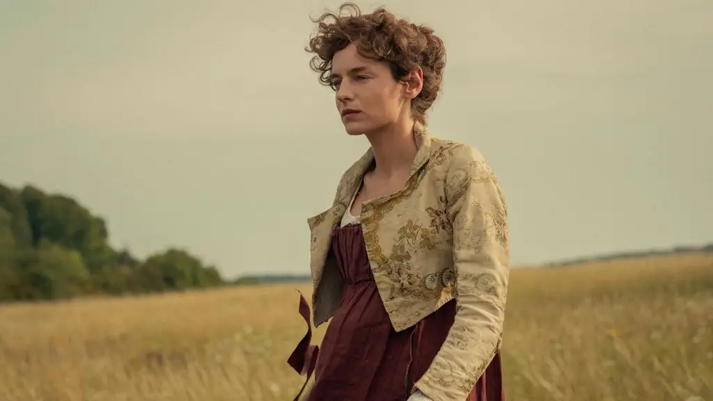 Emma Corrin as Elizabeth Bennet with curly brown hair stands in a field, wearing a red dress and beige jacket.