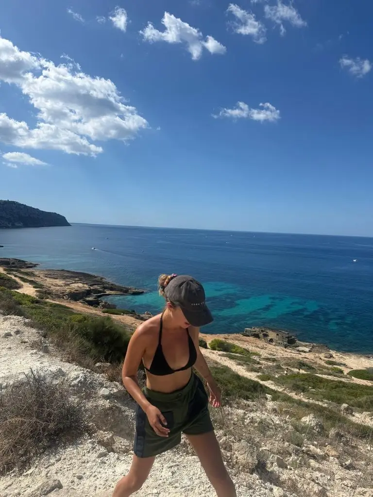 Duke of Kent's granddaughter Girl Amelia Windsor stuns in gorgeous Spanish bikini photographs 3 A woman dressed in a black bikini top and green shorts stands on a rocky beach, facing the ocean.