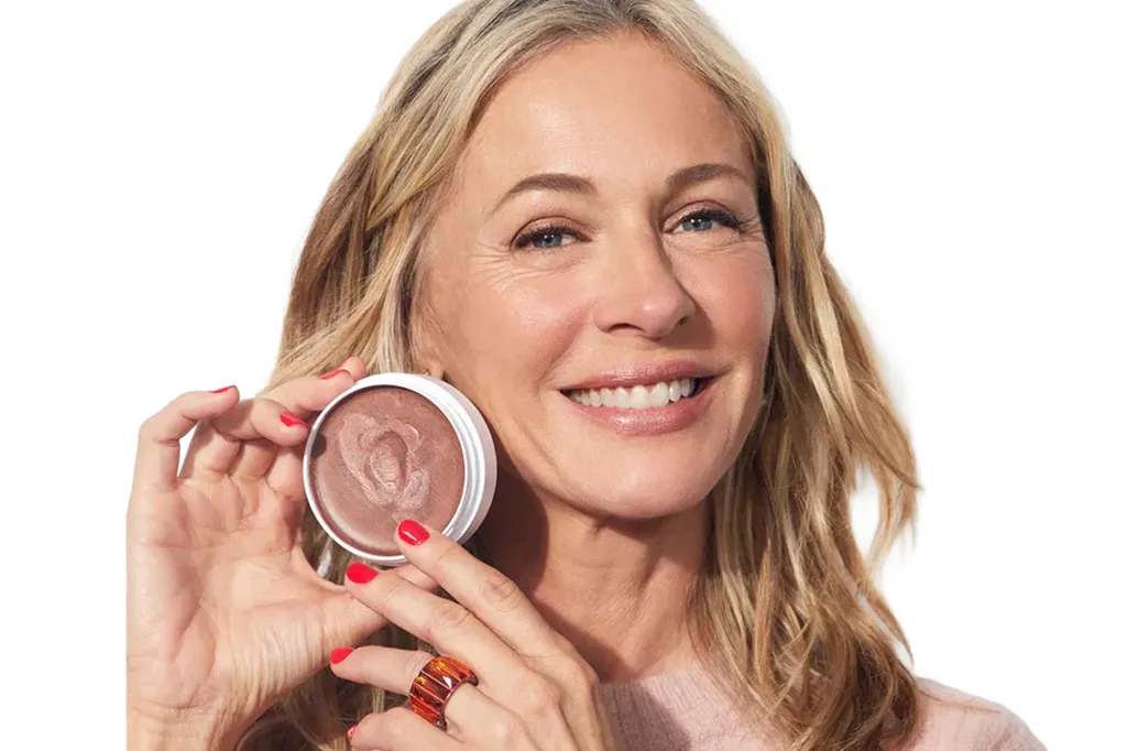 A woman with blonde hair, red nails and a light pink sweater smiles while holding a blush compact to her cheek.