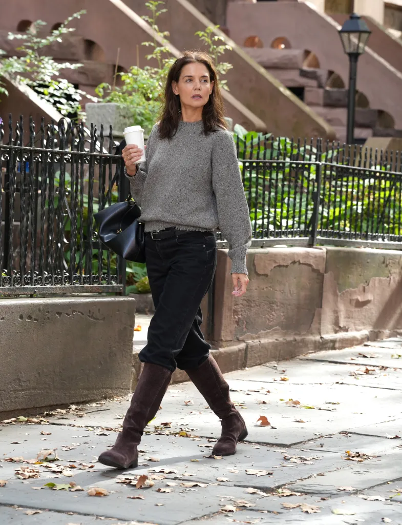 Katie Holmes walks on a sidewalk, holding a cup of coffee, wearing a gray sweater, black pants and brown boots.