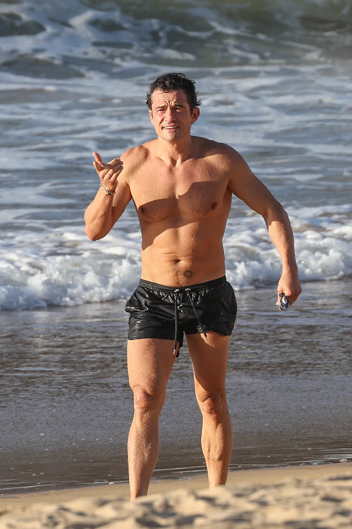 Orlando Bloom standing shirtless on La Concha beach, looking at the camera with an open-mouthed smile and left-hand gestures.