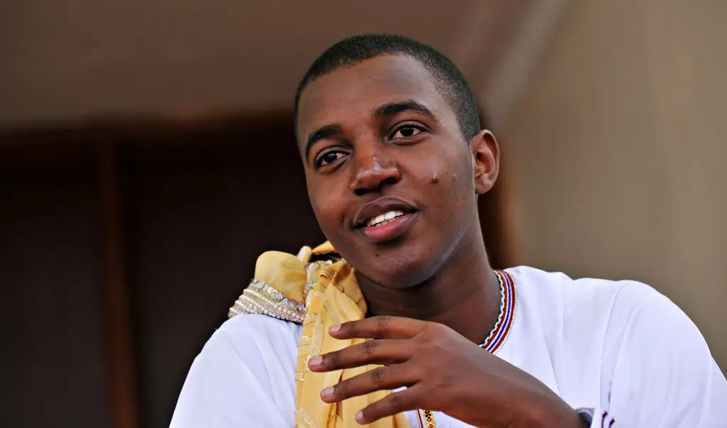 Person in white shirt and yellow scarf smiling and gesturing with one hand.