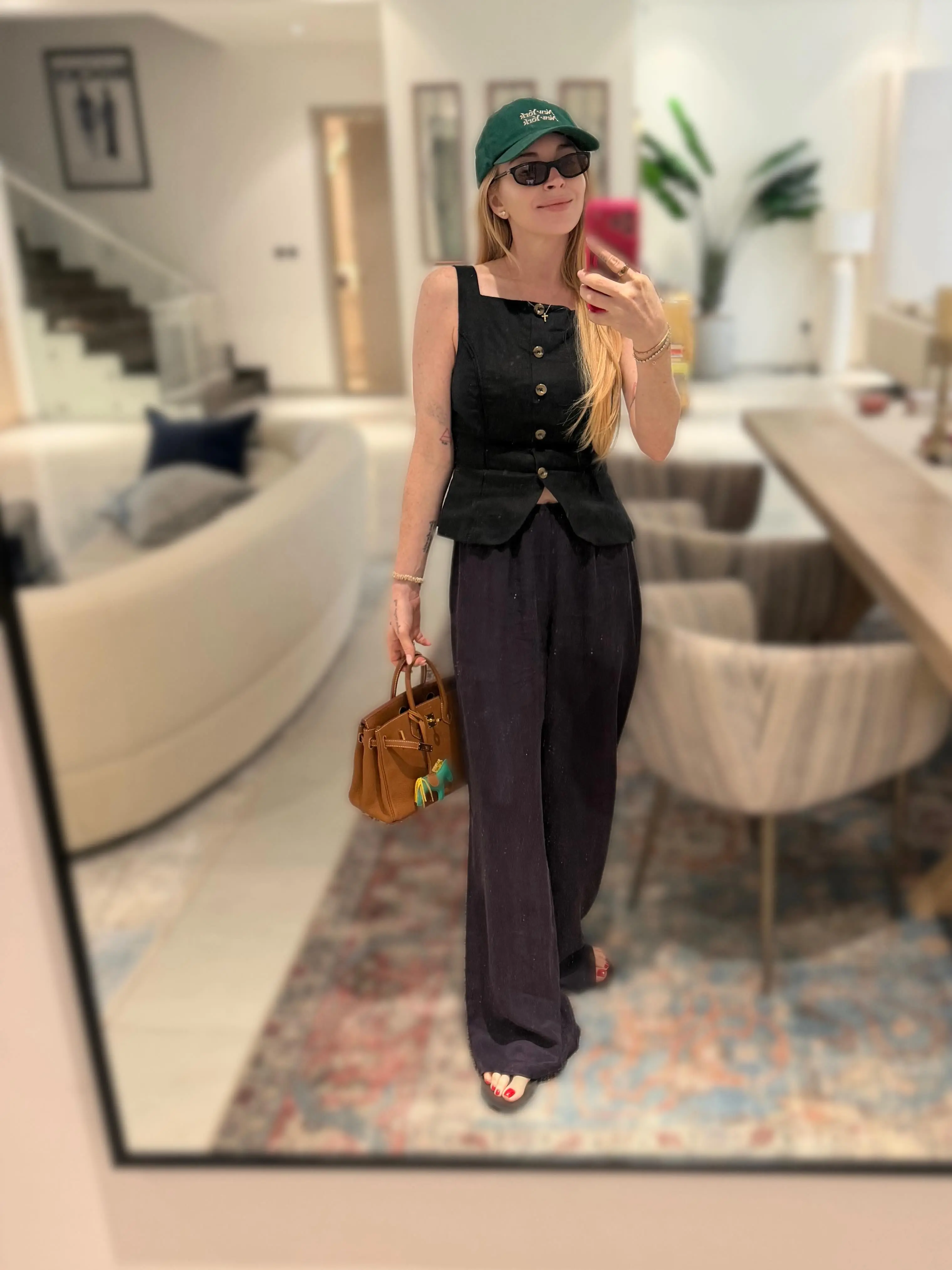 Lindsay Lohan wearing sunglasses, a dark green baseball cap, a black top and dark pants, holding a small brown purse.