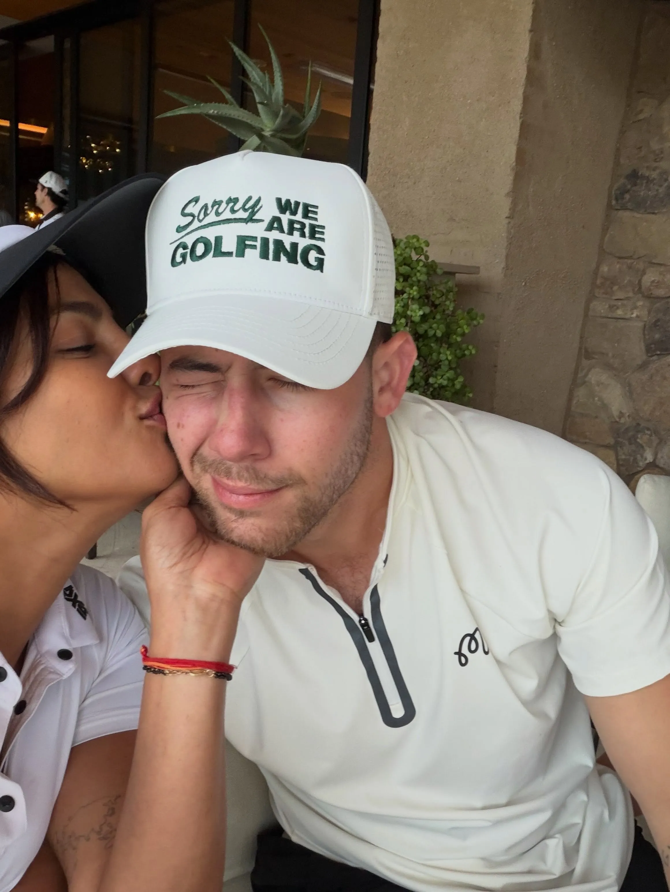 Priyanka Chopra kisses Nick Jonas on the cheek while he wears a "Sorry, we're playing golf" hat.