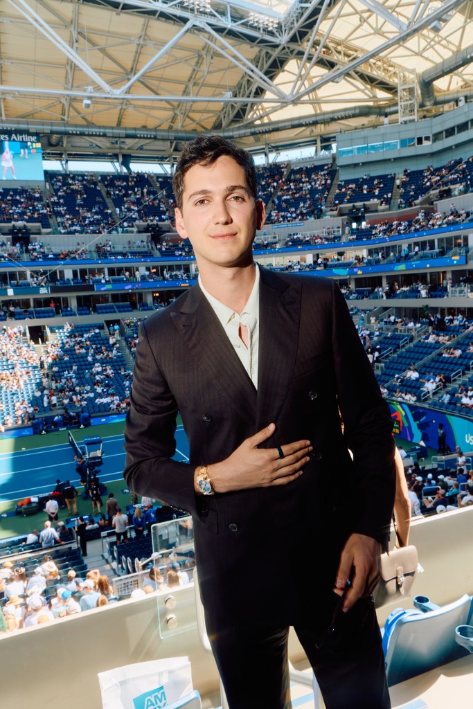 A man in a double-breasted suit smiles at the camera from a box at the US Open.