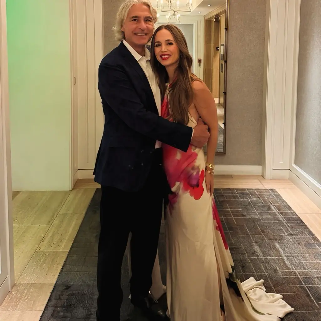Eliza Dushku, wearing a white and pink floral dress with her hair down, smiles as she is kissed by her husband, Peter Palandjian.