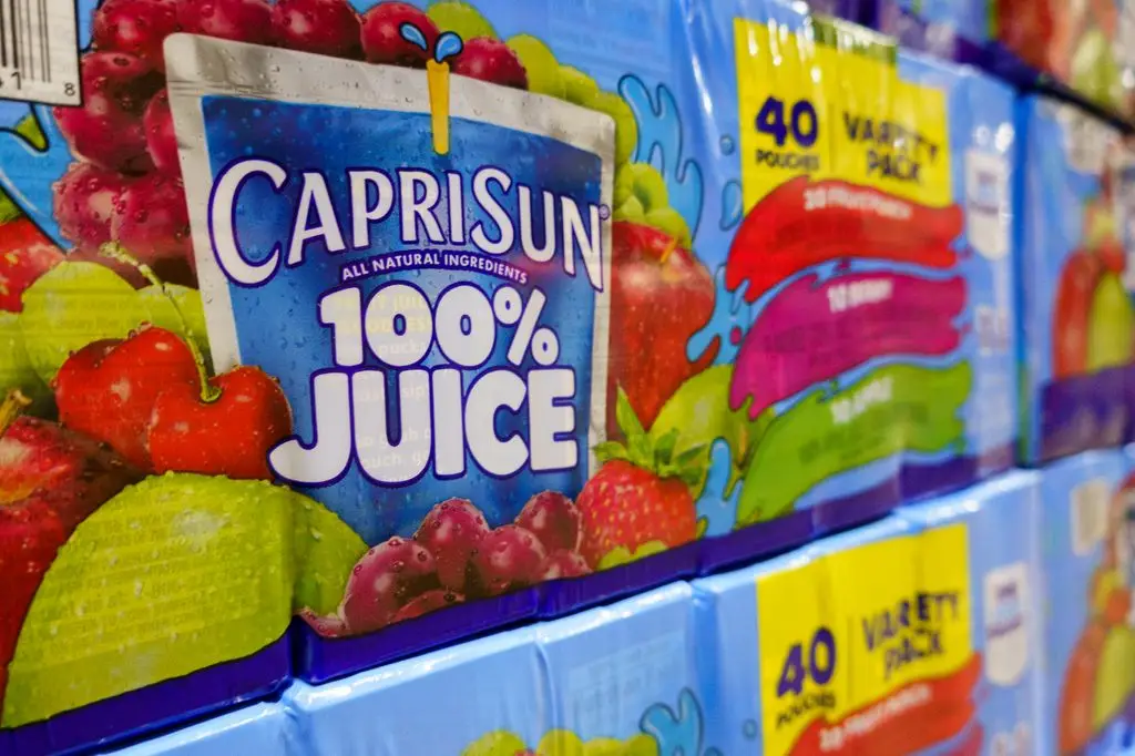 Close-up of packaging boxes of Capri Sun 100% juice varieties stacked on a store shelf.