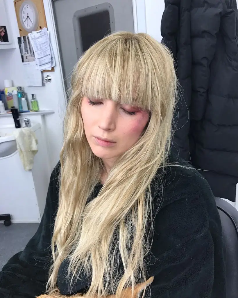 Jennifer Lawrence, wearing a dark dress, sits with her eyes closed as her long blonde hair with bangs is styled in a dressing room backstage.