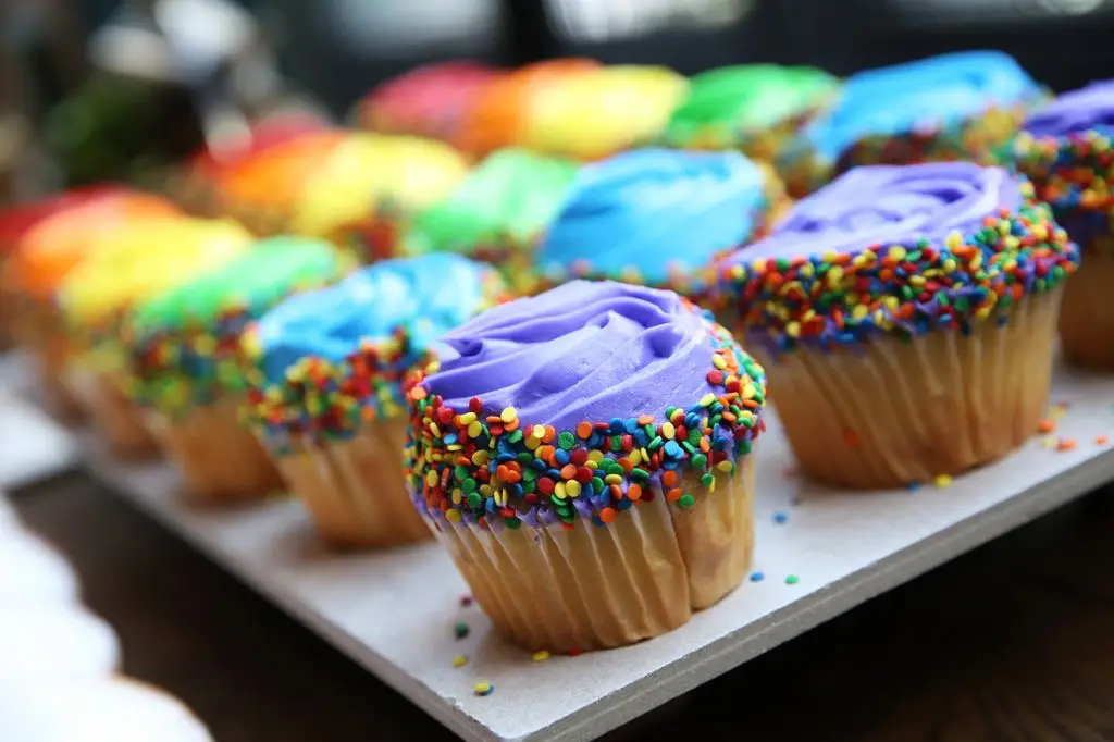 Rows of cupcakes with brightly colored frosting in rainbow tones, topped with multi-colored sprinkles, arranged on a platter.