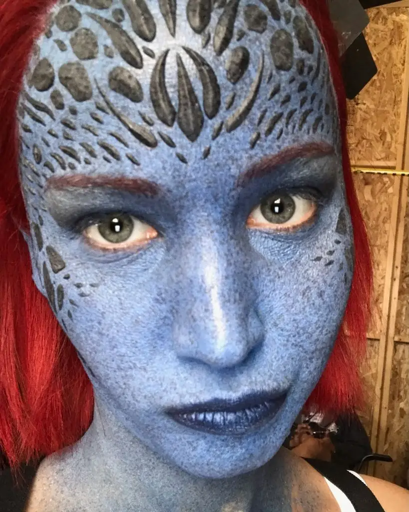 A close-up of Jennifer Lawrence in full Mystique makeup, with blue-scaled skin, dark reptilian patterns on her forehead, and red hair framing her face.