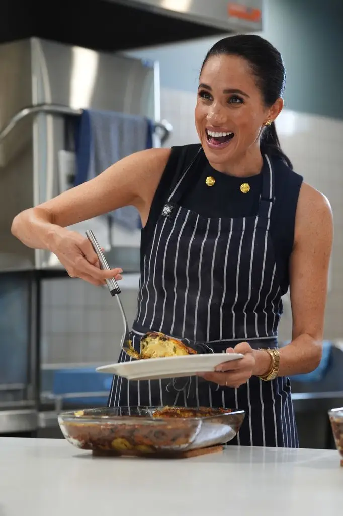 Meghan, Duchess of Sussex, wearing a black and white striped apron, holding a plate of food with a fork.