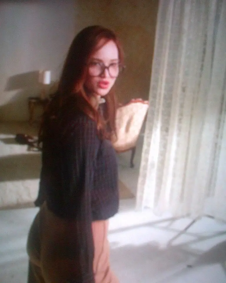 Jennifer Lawrence, wearing a red wig, glasses and a dark striped shirt, looks over her shoulder in a sunny room with lace curtains.
