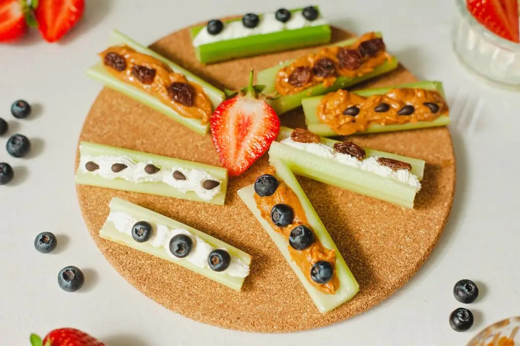 A cork tray topped with celery sticks filled with peanut butter or cream cheese and garnished with raisins, blueberries or chocolate chips, surrounded by fresh strawberries and blueberries.