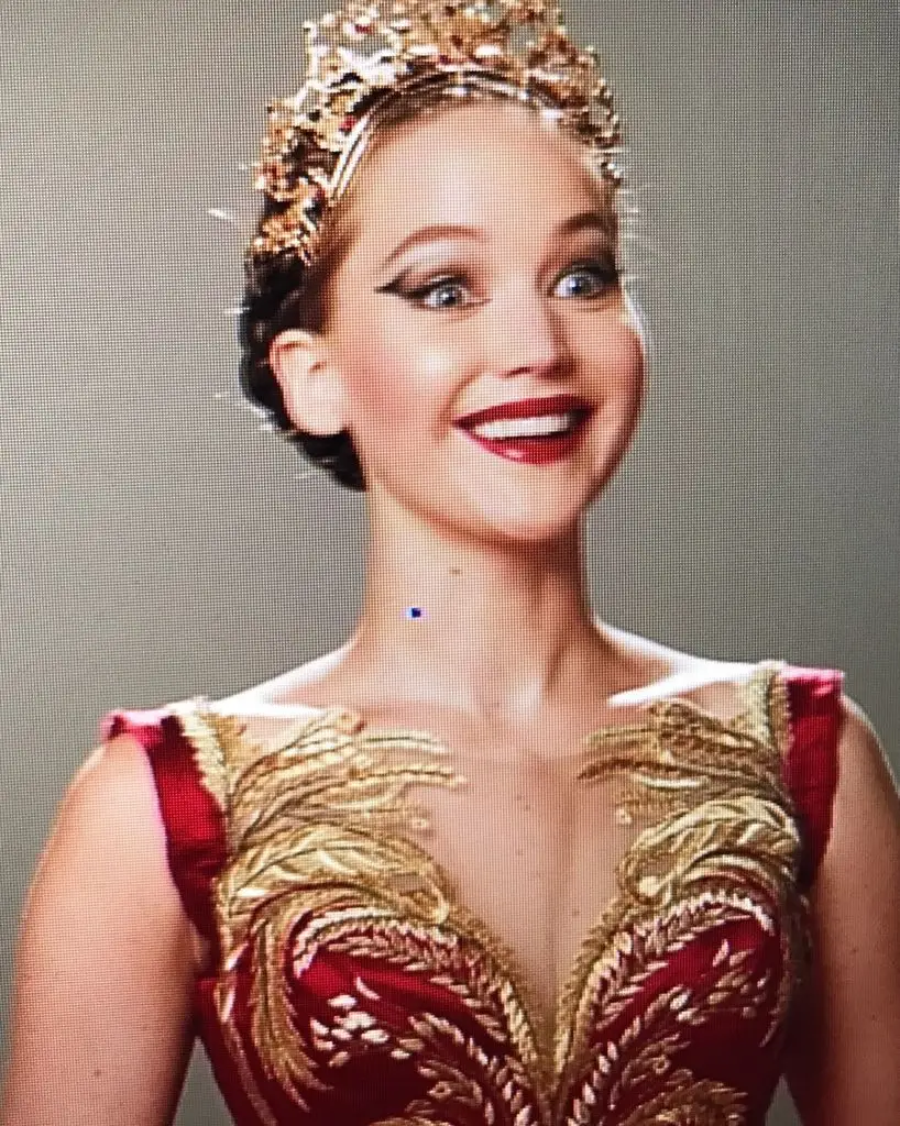 Jennifer Lawrence, wearing a gold crown and red and gold embroidered dress, smiles broadly with red lipstick.
