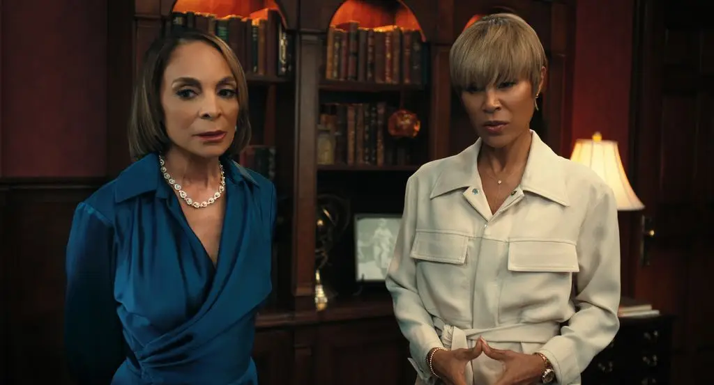 Jasmine Guy and Jada Pinkett Smith stand in a room with shelves and a lamp.