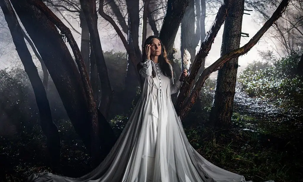 Carrie-Anne Moss stands in a foggy forest, holding a white mask and wearing a long white dress.