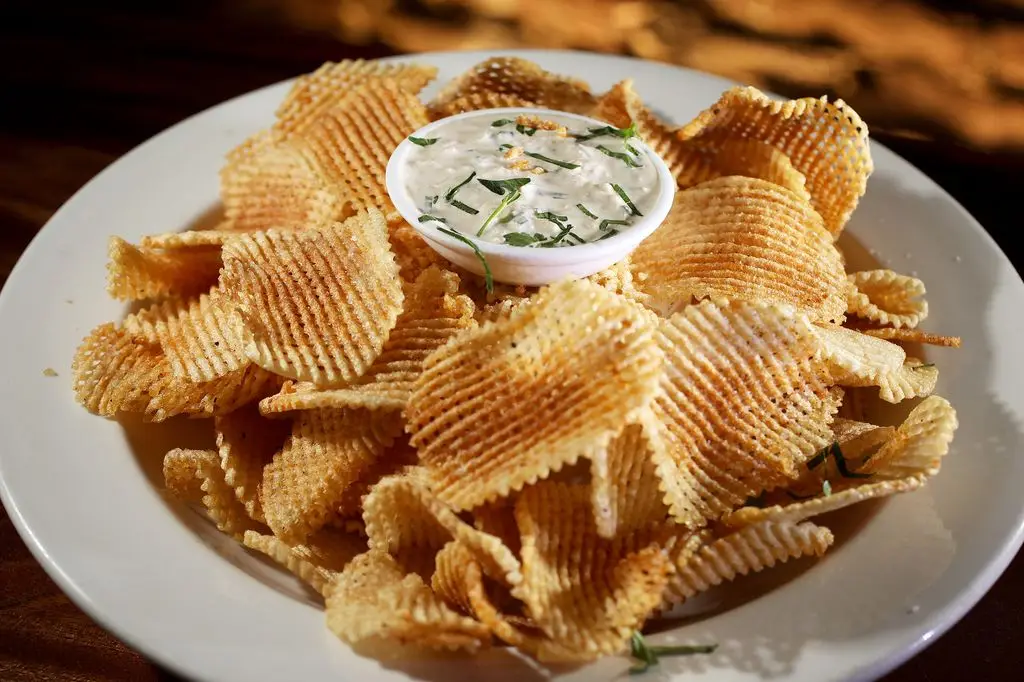 A white plate filled with wavy potato chips surrounding a small bowl of creamy onion dip topped with chopped herbs.
